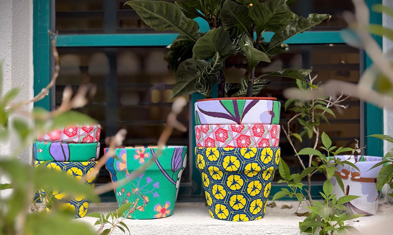 A row of fabric-covered terracotta pots in bold nala prints, stacked and planted, on a sunny windowsill.